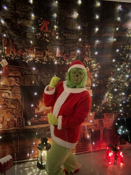 Grinch Grüner Charakter in rotem Weihnachtskostüm vor festlicher Hintergrunddekoration.